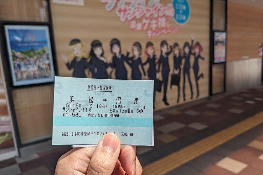 臨時急行 ラブライブ!サンシャイン!! 号の指定席券の写真。背景は沼津駅南口に掲示されているJR東海の制服を着たAqoursメンバー。
